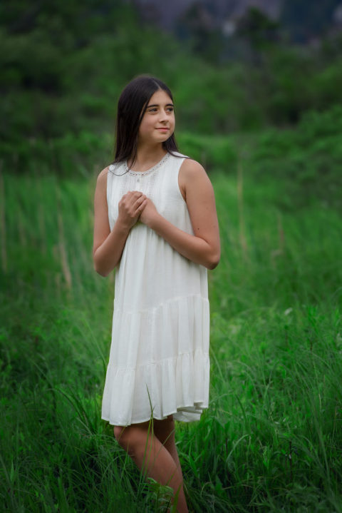 Family Portrait girl in white dress standing in green grass with hands on heart and looking to the right