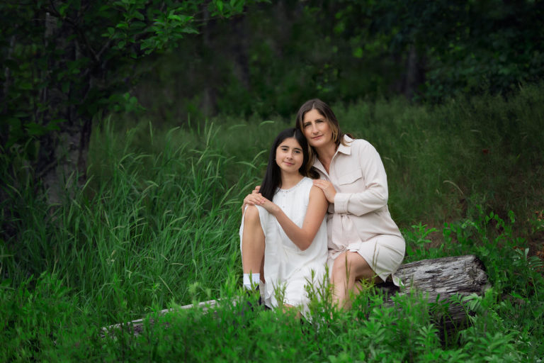 Family Portrait of Mom cuddling with daughter siting on log surrounded by green grass and small green shrubs