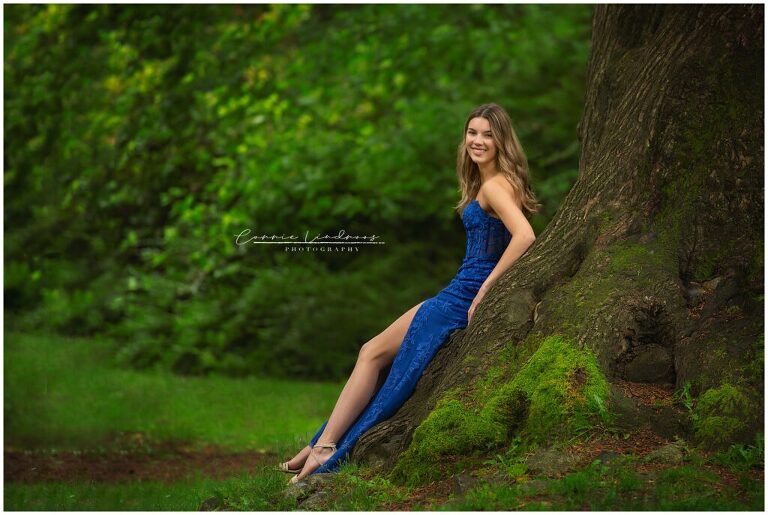 Female Highschool Graduate leaning on roots of tree, wearing royal blue prom dress