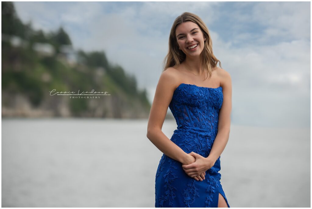 Female grad standing at the edge of the ocean with both hands on one hip wearing a royal blue prom dress.