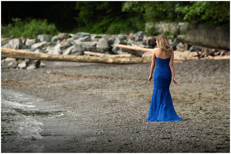 Female grad walking away from camera on the edge of a rocky beach, wearing a royal blue prom dress