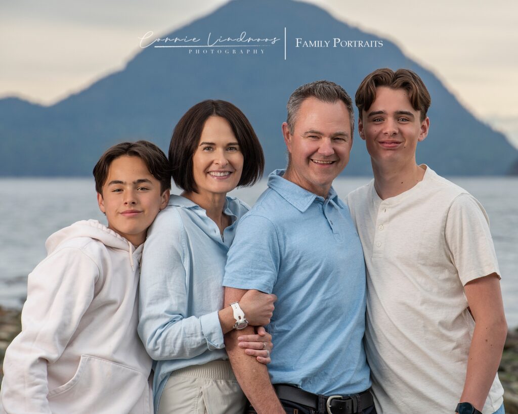 family photography mom, dad, two sons wearing blue and white, posed in front of the ocean with the mountains behind them